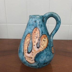Pode incluir: Um pequeno jarro de cerâmica azul com uma pega. O jarro apresenta um desenho pintado à mão de uma forma semelhante a um cacto em bege. O jarro tem aproximadamente 13 cm de altura e é adequado para uso decorativo.