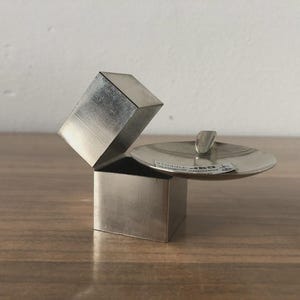Peut inclure: Petite boîte argentée avec un couvercle articulé, ouvert pour révéler l'intérieur. Le couvercle est de forme plate et circulaire avec une petite poignée. La boîte est posée sur une surface en bois.
