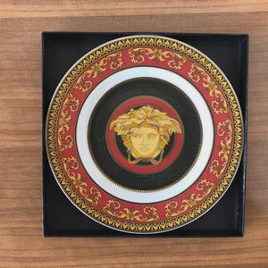 Pode incluir: Prato decorativo com um centro preto com um design de cabeça de Medusa dourada. O prato tem uma borda vermelha e dourada ornamentada e um anel interno branco. O prato é apresentado em uma caixa preta.