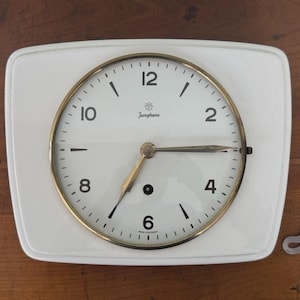 May include: A white wall clock with a rounded rectangular frame and a circular face featuring a gold-toned bezel. The clock face displays black Arabic numerals, the Junghans logo, and metallic hands. A separate metal winding key is included.