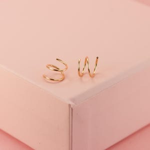 May include: A pair of gold-coloured spiral earrings. One earring features a double spiral, the other a triple spiral design. The earrings are presented on a light pink surface.