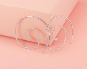 May include: A pair of silver spiral earrings. The earrings are displayed on a light pink surface, with a matching pink background. The earrings have a minimalist design, with a curved, open spiral shape.