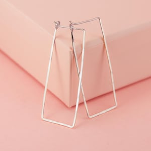 May include: A pair of silver rectangular hoop earrings. The earrings are set against a soft pink backdrop. The earrings have a hammered texture and a simple, modern design, suitable for everyday wear.