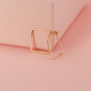 Könnte beinhalten: Ein Paar goldfarbene quadratische Creolen. Die Ohrringe werden auf einer hellrosa Oberfläche mit einer passenden rosa Schachtel im Hintergrund präsentiert. Die Ohrringe haben ein schlichtes, geometrisches Design.