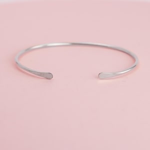 Puede incluir: Un brazalete abierto de color plateado. El brazalete tiene un diseño simple y minimalista con extremos redondeados. El fondo es de color rosa claro liso.
