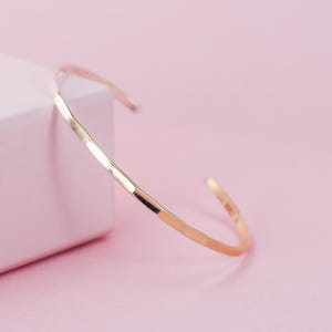 Puede incluir: Un brazalete de puño de color dorado con un acabado martillado. El brazalete abierto tiene un diseño simple y elegante. El brazalete se muestra sobre un fondo rosa junto a una superficie blanca.