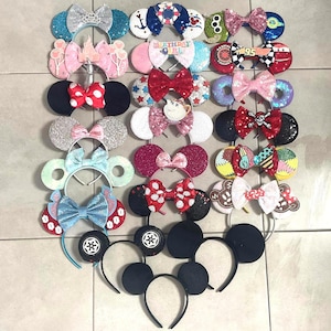 Puede incluir: Una colección de veintitrés diademas decorativas con orejas circulares y varios lazos temáticos. Los diseños incluyen texturas brillantes, patrones inspirados en personajes, lentejuelas y motivos específicos como un pastel de cumpleaños.