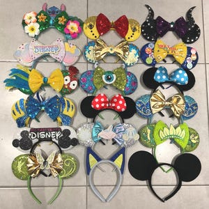 Puede incluir: Una colección de diademas decorativas con varios diseños. Algunas presentan orejas de ratón, lazos y adornos temáticos de personajes. Los colores incluyen rojo, amarillo, azul y verde, con acentos de purpurina y lentejuelas. Algunas diademas tienen el texto "My First Trip to Disney".