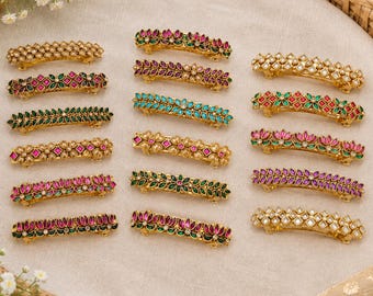 May include: A collection of fifteen gold-tone metal hair barrettes arranged in three columns. Each barrette features intricate floral or geometric patterns inlaid with colorful stones in shades of pink, green, blue, and clear white crystals.