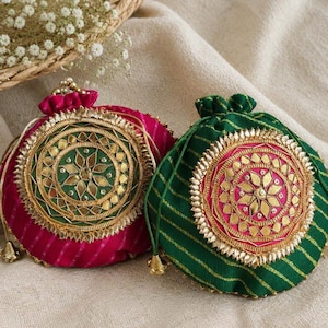 Puede incluir: Dos bolsas con cordón, una magenta y otra verde, ambas con un medallón central circular con bordados dorados, detalles tipo espejo y un patrón de tela a rayas. Cada bolsa tiene una parte superior fruncida con cuentas de metal en tono dorado.