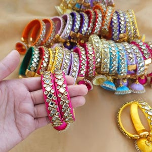 May include: A collection of vibrant, silk-wrapped bangles in various colors including pink, yellow, blue, purple, and orange. Each bangle features a central row of square and circular mirrors set in gold-toned metal frames, with some including small dangling charms.