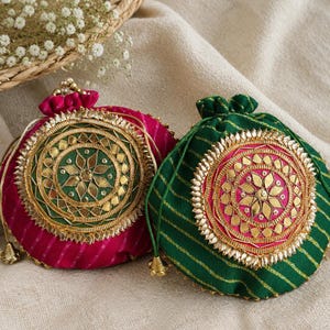 May include: Two drawstring pouches featuring a circular central medallion with gold-toned embroidery and mirror accents. One pouch is magenta with diagonal stripes, and the other is emerald green with diagonal stripes and a pink center medallion.