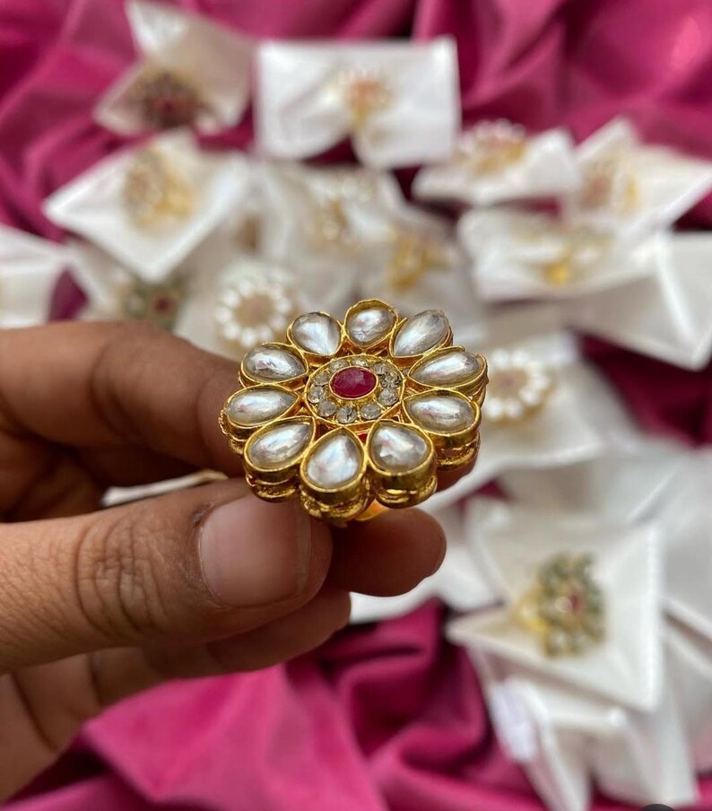 May include: A gold-toned ring with a floral design. The ring features a central red gemstone surrounded by smaller clear stones. The petals are pear-shaped, with a white, pearl-like appearance. The ring is held against a backdrop of other jewelry.