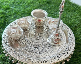 May include: Ornate silver puja thali set with a central swastika design. The set includes a large, round tray with floral patterns, small bowls, a cup, and a bell-shaped item with a handle. The tray is decorated with dangling charms.