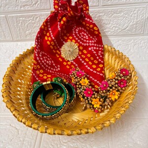 Könnte beinhalten: Ein geflochtener Korb enthält einen roten Kordelzugbeutel mit einem gelb-weißen Muster, eine goldene Blumenverzierung, grüne Armreifen und eine Blumenkette mit rosa und gelben Blüten. Der Korb ist aus geflochtenem Bambus.