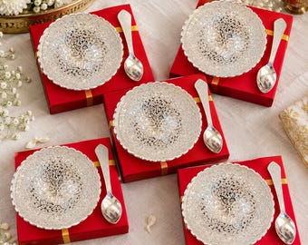 May include: Five silver-colored bowls with intricate floral cut-out patterns, each paired with a matching silver-colored spoon. Each set is presented on a square red gift box, arranged on a light-colored fabric surface with white flowers.