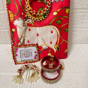 May include: A red fabric tote bag with a white cow illustration, surrounded by floral patterns. The bag is accompanied by a framed set of bindi, hair accessories, and red and gold bangles. A decorative handle is attached to the bag.
