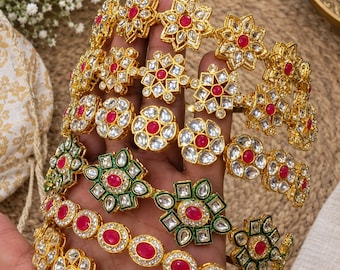 May include: A set of gold-toned bangles featuring floral motifs with embedded clear stones and central pink cabochons. Some bangles include green enamel detailing around the floral patterns, while others maintain a solid gold-toned metal finish.