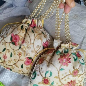 Könnte beinhalten: Zwei cremefarbene Beutel mit Kordelzug und floraler Stickerei in Rosa, Rot und Grün. Die Beutel haben goldfarbene Akzente und eine Perlenbordüre. Jeder Beutel hat einen perlen- und goldperlenbesetzten Griff.