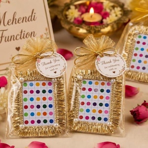 Könnte beinhalten: Zwei rechteckige Gastgeschenke in transparenten Organzabeuteln mit goldenen Schleifen. Jedes Geschenk hat einen goldfarbenen Rahmen mit einem Gitter aus bunten Bindi-Punkten. Ein weißes, rundes Etikett trägt die Aufschrift Thank You for celebrating with us.