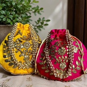 Könnte beinhalten: Zwei Beutel mit Kordelzug, einer in leuchtendem Gelb und einer in kräftigem Pink, mit aufwendigen goldenen Blumenstickereien und reflektierenden Details. Jede Tasche hat einen langen, mehrsträngigen Tragegriff aus weißen Perlen und einen gerafften Verschluss.