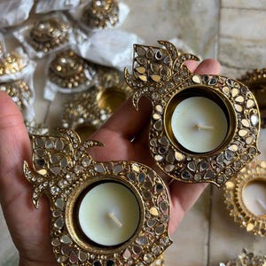 May include: Two gold-toned metal tea light candle holders shaped like pomegranates, featuring intricate filigree patterns and embedded reflective mirror pieces. Each holder contains a small, round, white wax candle with a centered wick.