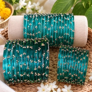 May include: A set of translucent teal glass bangles featuring small, metallic silver-toned floral accents distributed across the surface. The bangles are arranged in three distinct groupings, showcasing a smooth, rounded profile and a vibrant, deep green-blue colour.