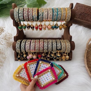 May include: A collection of multi-colored bangles featuring floral stone embellishments and small hanging charms, displayed on a two-tiered wooden stand, accompanied by a set of square coasters with colorful fringe and polka dot patterns.