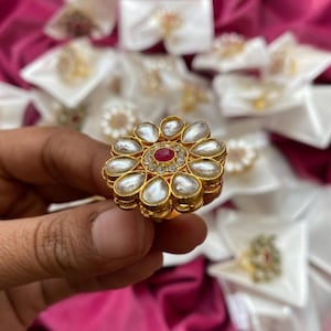 May include: A gold-toned ring with a floral design. The ring features a central red gemstone surrounded by smaller clear stones. The petals are pear-shaped, with a white, pearl-like appearance. The ring is held against a backdrop of other jewelry.