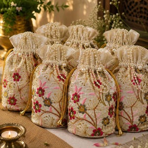 May include: A collection of cream-colored drawstring pouches featuring intricate floral embroidery in shades of pink, red, and green. Each pouch is cinched with a pearl-beaded drawstring and finished with gold-toned metal tassels at the ends.
