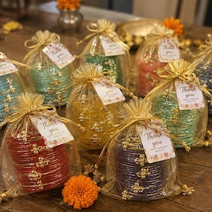 Puede incluir: Una colección de nueve bolsas de organza dorada transparente con cordón, cada una con una pila de brazaletes finos y brillantes en varios colores, incluyendo rojo, amarillo, verde, azul y morado. Cada bolsa tiene una etiqueta con Thank you.