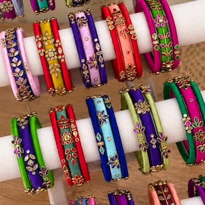 May include: A collection of multi-colored silk thread bangles arranged on a white display stand. Each bangle features intricate metallic embellishments, including floral patterns and geometric shapes set with small, reflective stones in various colors.