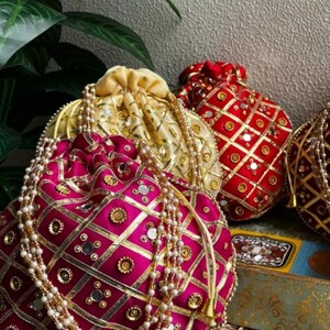 May include: A selection of decorative drawstring bags in shades of fuchsia, gold, and red. The bags are adorned with gold trim, small mirrors, and pearl-like beads. The bags are a traditional design.