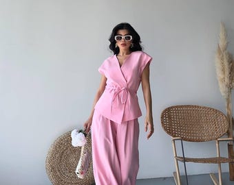 May include: A light pink two-piece outfit consisting of a wrap top and wide-leg pants. The top has short sleeves and a tie closure. The outfit is paired with white-framed sunglasses and a woven bag.