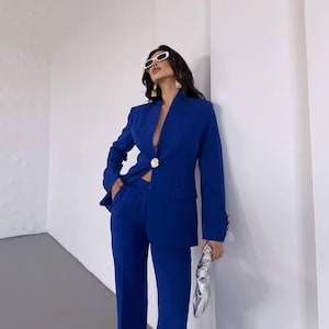 Puede incluir: Un traje azul vibrante, con una chaqueta con botón dorado y pantalones a juego. El conjunto se complementa con gafas de sol blancas y un clutch plateado. El diseño del traje es elegante y moderno, perfecto para un entorno profesional o formal.