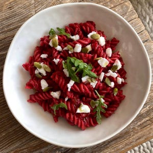 May include: A white bowl filled with red pasta, garnished with white cheese, green arugula, and pumpkin seeds. The pasta is a vibrant red color, and the dish is set on a wooden surface, creating a rustic aesthetic.