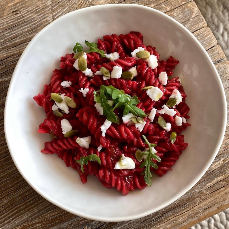 May include: A white bowl filled with red pasta, garnished with white cheese, green arugula, and pumpkin seeds. The pasta is a vibrant red color, and the dish is set on a wooden surface, creating a rustic aesthetic.
