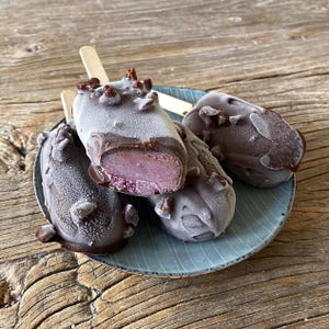 May include: Four frozen ice cream bars on a blue plate. The bars are rectangular, with a dark chocolate coating and a light purple interior. Some bars have a bite taken out, revealing the purple ice cream. The plate sits on a wooden surface.