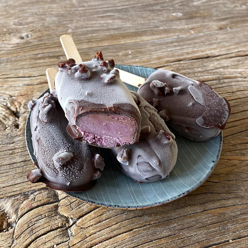 May include: Four frozen ice cream bars on a blue plate. The bars are rectangular, with a dark chocolate coating and a light purple interior. Some bars have a bite taken out, revealing the purple ice cream. The plate sits on a wooden surface.