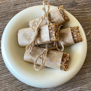 May include: A light-colored ceramic plate holds several wrapped food bars. Each bar is wrapped in brown paper and tied with twine. The bars have a textured, granular appearance, with visible seeds and grains. The plate sits on a wooden surface.