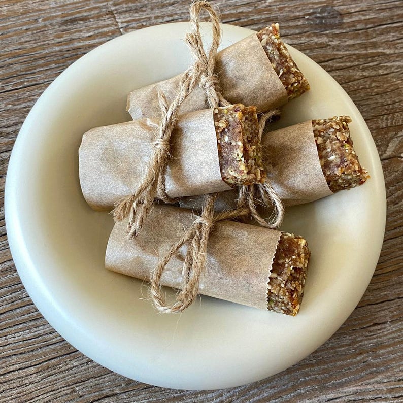 May include: A light-colored ceramic plate holds several wrapped food bars. Each bar is wrapped in brown paper and tied with twine. The bars have a textured, granular appearance, with visible seeds and grains. The plate sits on a wooden surface.