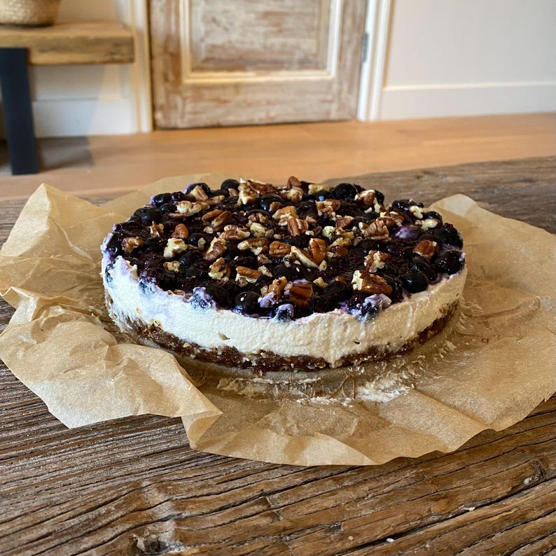 May include: A round cheesecake with a graham cracker crust, a creamy white filling, and a topping of blueberries and chopped pecans. The cake sits on a sheet of brown parchment paper on a wooden surface.
