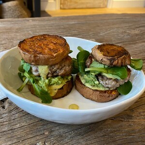 May include: Two mini burgers with sweet potato buns, meat patties, avocado spread, and greens, served in a white bowl. The sweet potato buns are grilled and golden brown. The avocado spread is a vibrant green.