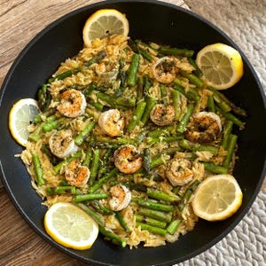 May include: A top-down view of a black skillet filled with a shrimp and asparagus orzo dish. The dish is garnished with lemon slices. The shrimp are seasoned and cooked. The asparagus is bright green. The orzo is a light brown color.