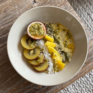 May include: A beige bowl filled with a colorful breakfast. Sliced golden kiwi, passion fruit, and mango are arranged around a creamy base topped with seeds and coconut flakes. The bowl sits on a wooden surface.