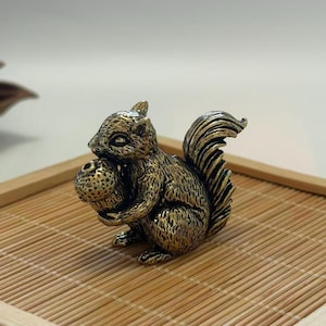 May include: A detailed bronze squirrel figurine holding an acorn. The squirrel has textured fur and a bushy tail. It sits on a bamboo tray with a light-colored background. The figurine is a decorative item.