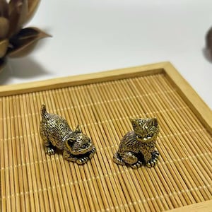 Könnte beinhalten: Zwei kleine, detaillierte Katzenfiguren aus Messing auf einem Bambustablett. Eine Katze liegt, die andere sitzt. Die Katzen haben strukturiertes Fell und sind goldfarben. Das Tablett ist quadratisch mit einem hellen Holzrahmen.