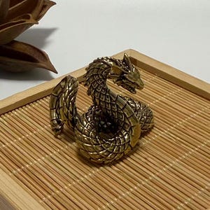 Puede incluir: Una detallada escultura de latón de una serpiente enroscada con intrincados detalles de escamas y cabeza. La serpiente está colocada sobre una bandeja de bambú con un marco de madera, y una flor seca en el fondo. La escultura tiene un acabado dorado metálico.
