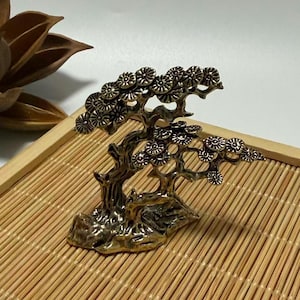 May include: A bronze-colored miniature tree sculpture with detailed branches and circular leaf patterns. The sculpture is mounted on a small base and sits on a bamboo tray, suggesting a decorative or ornamental purpose.