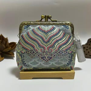 May include: A small, ornate coin purse with a clasp closure. The purse features a detailed, patterned fabric in shades of blue, red, and gold. It has a decorative metal frame and a small tassel. The purse is displayed on a small wooden stand.